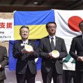 福井県庁前で開かれた出発式で、パックご飯を手に写真に納まる杉本達治知事（左から2人目）ら＝25日午前